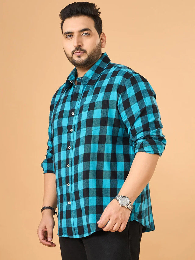 Plus size man in blue and black checked shirt with rolled sleeves, black jeans, neutral background.
