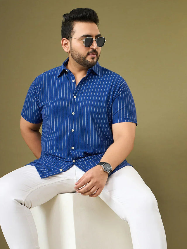 Plus size man wearing a blue striped short sleeve shirt and white pants, seated, beige background