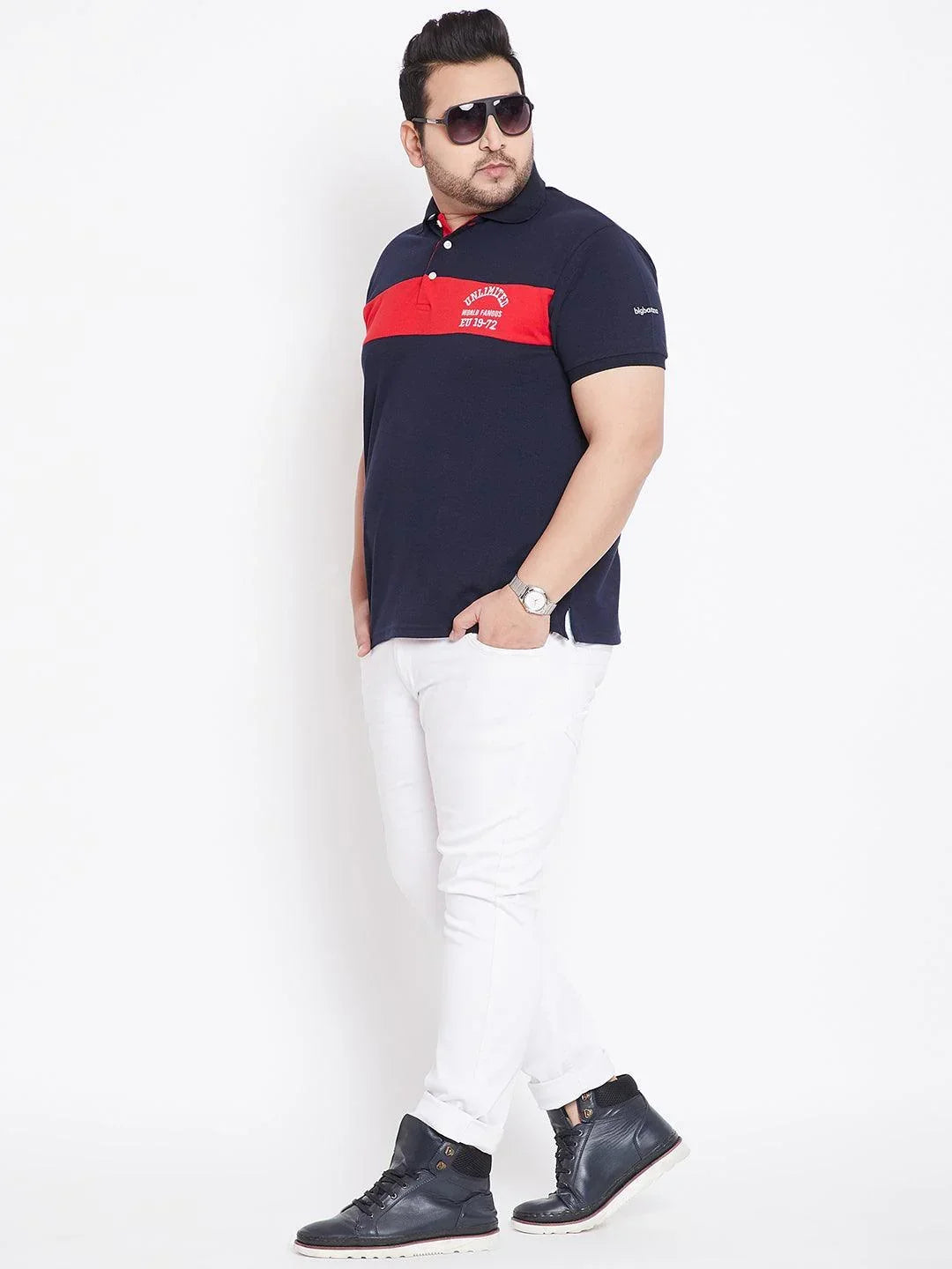 Plus size man in navy polo shirt with red stripe, white pants, black boots and sunglasses
