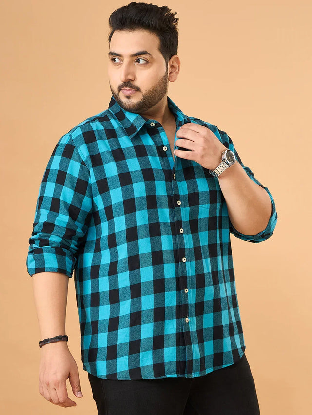 Plus size man in teal and black checkered shirt with rolled sleeves, posing against tan background