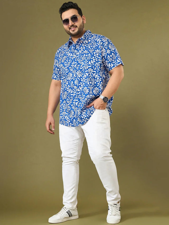 Plus size man in blue floral short sleeve shirt, white jeans, and white sneakers posing confidently.